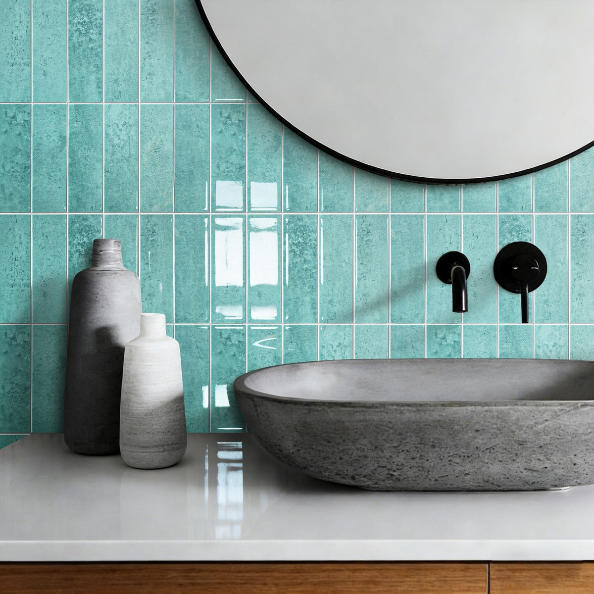 A modern bathroom scene featuring glossy turquoise rectangular wall tiles, a grey stone vessel sink on a white countertop, two grey decorative bottles, and a round mirror.