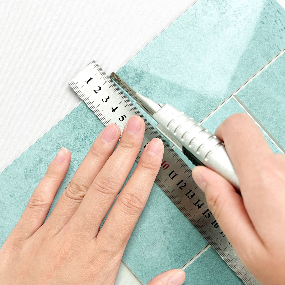 A close-up of a person's hands using a silver utility knife and metal ruler to measure and cut a light blue-green textured tile sheet.