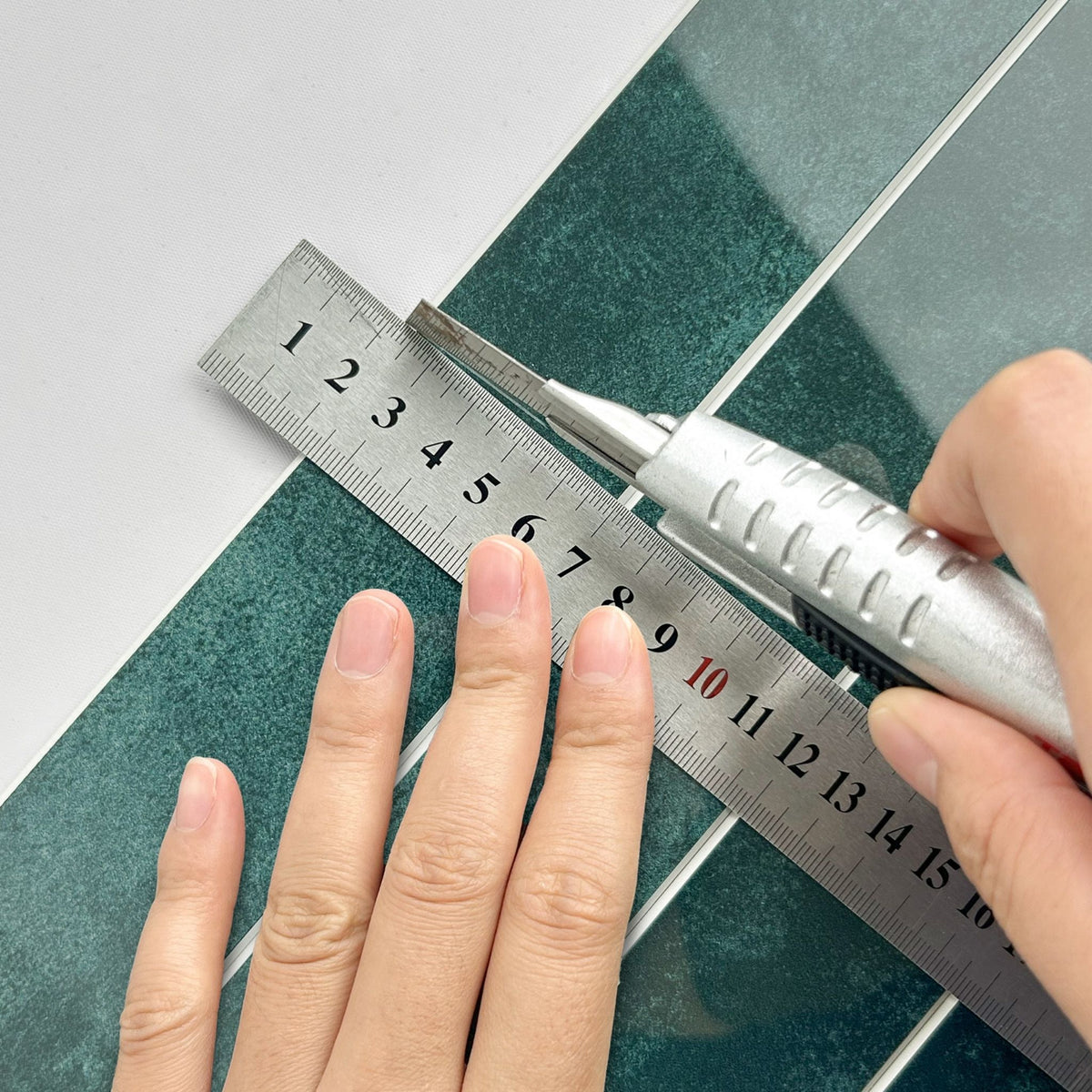 A close-up of a hand using a silver metal ruler and utility knife to measure and cut a dark green composite tile, on a white background.