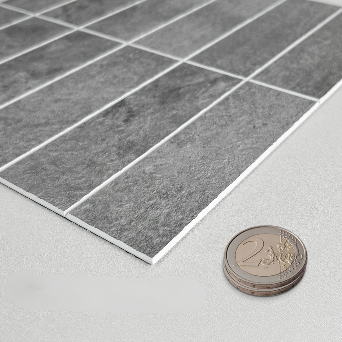 Grey rectangular wall tiles with white grout lines, shown at an angle on a light surface with a 2 Euro coin for scale.