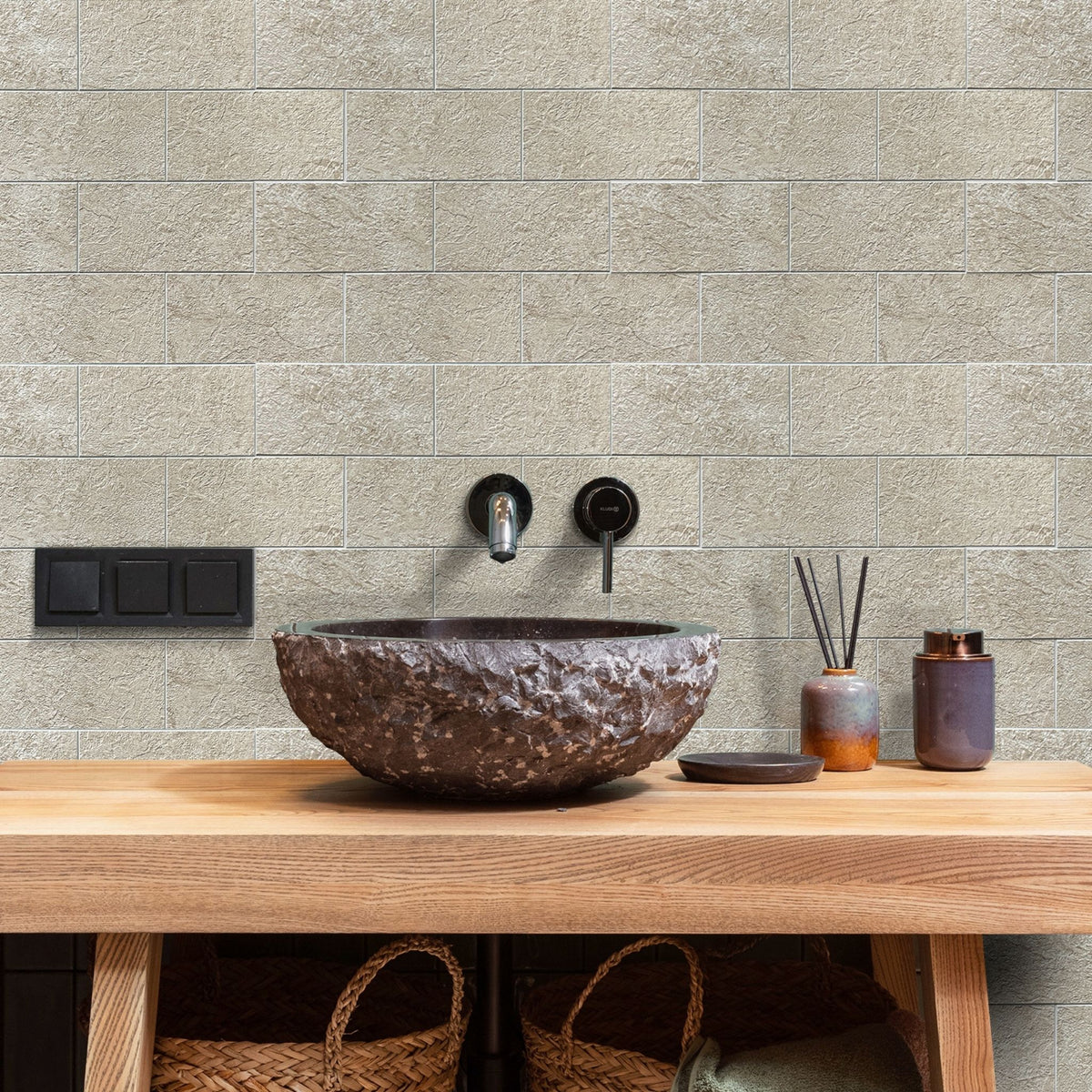A bathroom vanity setup with a large textured dark brown stone vessel sink on a light wood countertop, set against a beige stone tile backsplash and black wall-mounted fixtures.