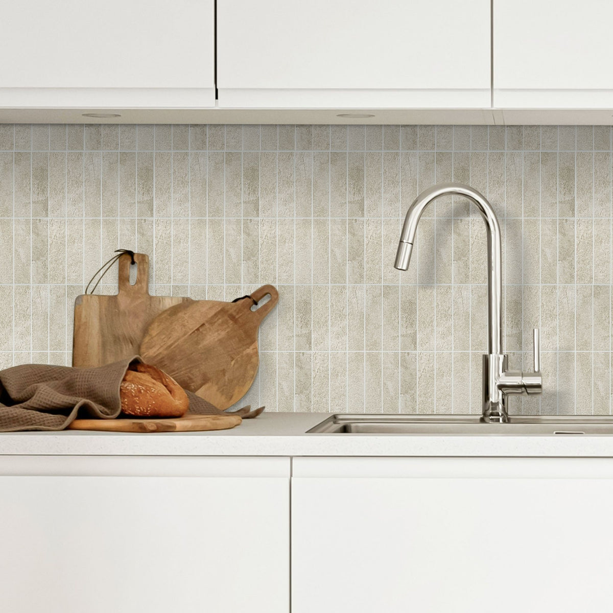 Stacked beige subway tiles as a backsplash in a white kitchen with a tap and wooden cutting boards and a loaf of bred on the counter.