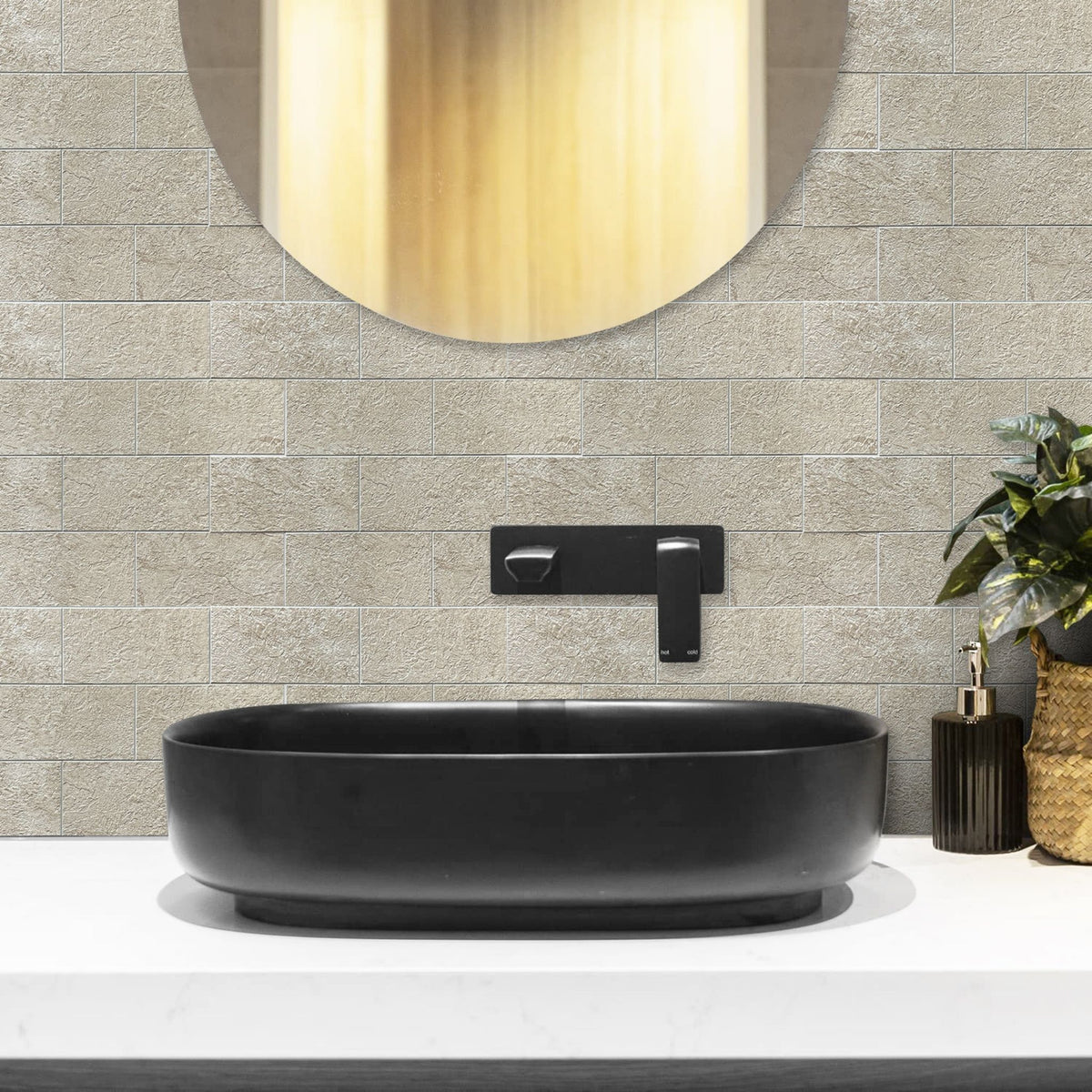 A modern bathroom scene featuring a matte black oval vessel sink mounted on a white countertop, set against a light beige stone tile backsplash with a matching black wall-mounted faucet. A round mirror and a small potted plant are also visible.
