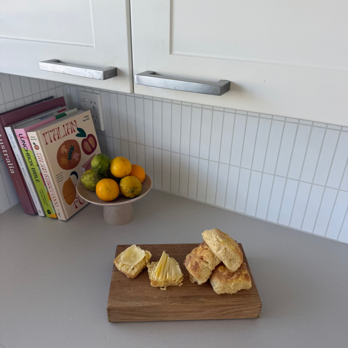 A wooden cutting board with sliced cheese and scones on a kitchen counter, with a fruit bowl and cookbooks in the background with white kit kat tiles.